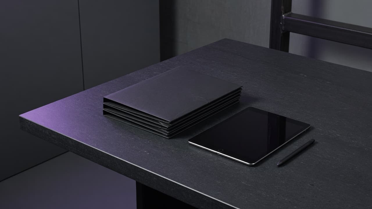 Dark press media table with plain matte black folders, blank tablet, and stylus under cool violet edge light.
